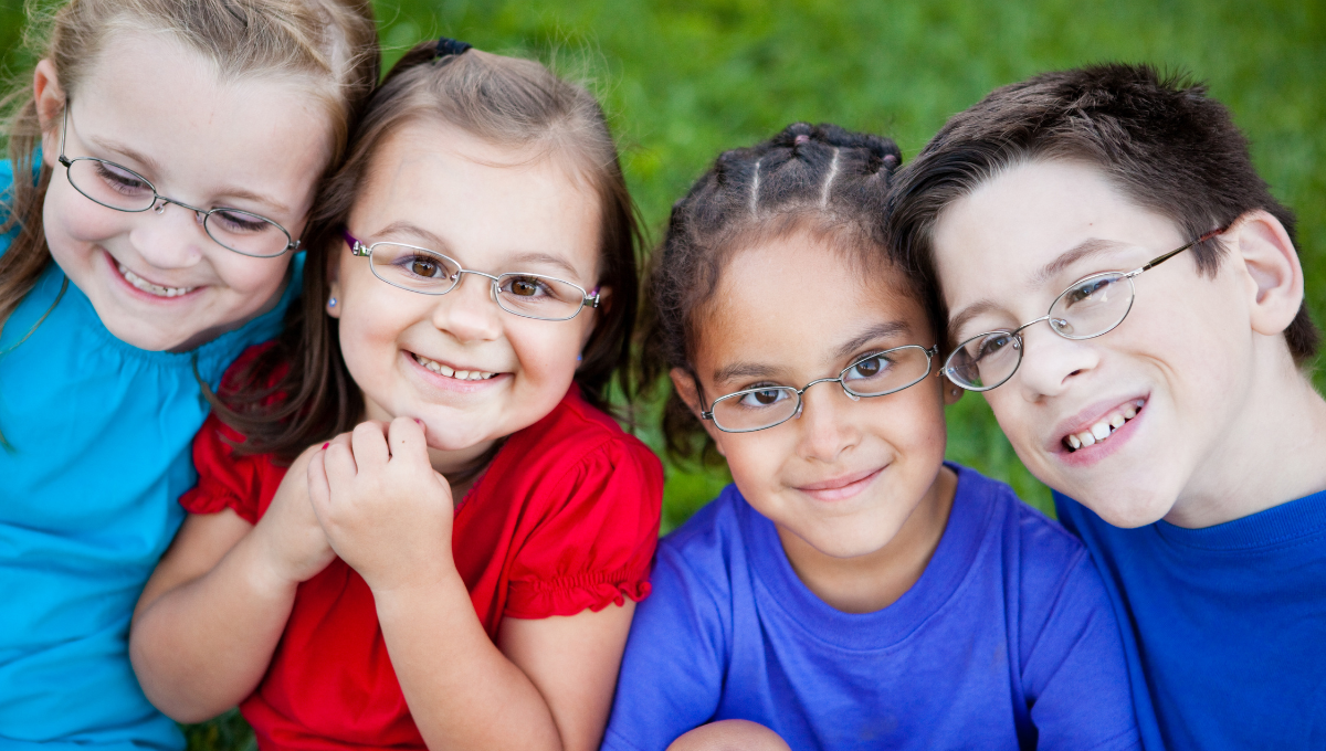 examen de la vue enfant Quatre enfants souriants portant des lunettes après un examen de la vue, assis sur l’herbe