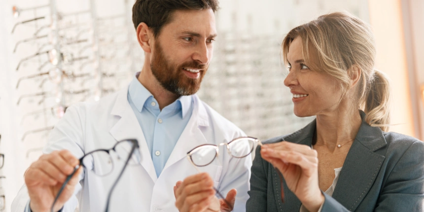 Deux professionnels de la santé visuelle, en blouse blanche, souriant devant un mur d'étagères à lunettes dans une clinique d'optique